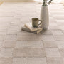 A light beige woven area rug with a textured block pattern, styled in a cozy room setting with a stack of books, a green ceramic mug, and a white textured vase on top.