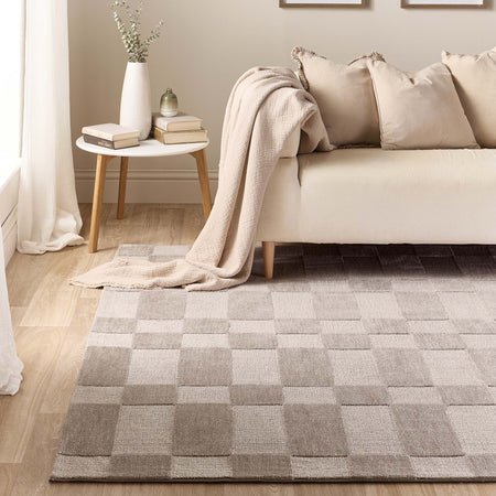 A rectangular area rug with a large-scale checkerboard pattern in neutral shades of taupe, beige, and light gray, styled in a modern living room setting next to a cream sofa and a small side table.