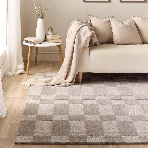 A rectangular area rug with a large-scale checkerboard pattern in neutral shades of taupe, beige, and light gray, styled in a modern living room setting next to a cream sofa and a small side table.