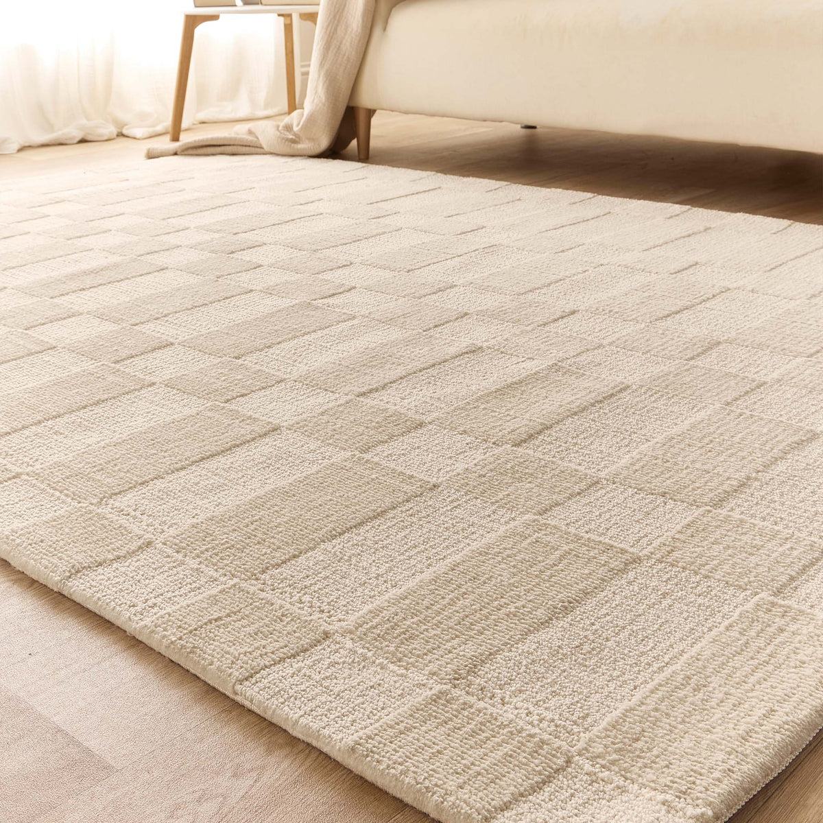 A light beige textured area rug with a woven geometric block pattern, shown in a modern bedroom setting on a light wood floor next to a bed.