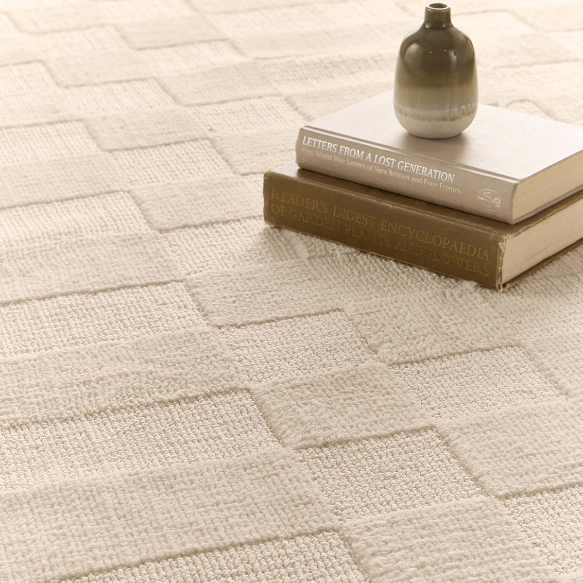 A close-up of a textured cream-colored area rug with a geometric basketweave pattern, styled with a small vase and two vintage books.