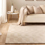 A large woven area rug in a warm cream and beige tone, styled in a cozy living room setting under a light beige sofa with matching pillows and a draped throw blanket.