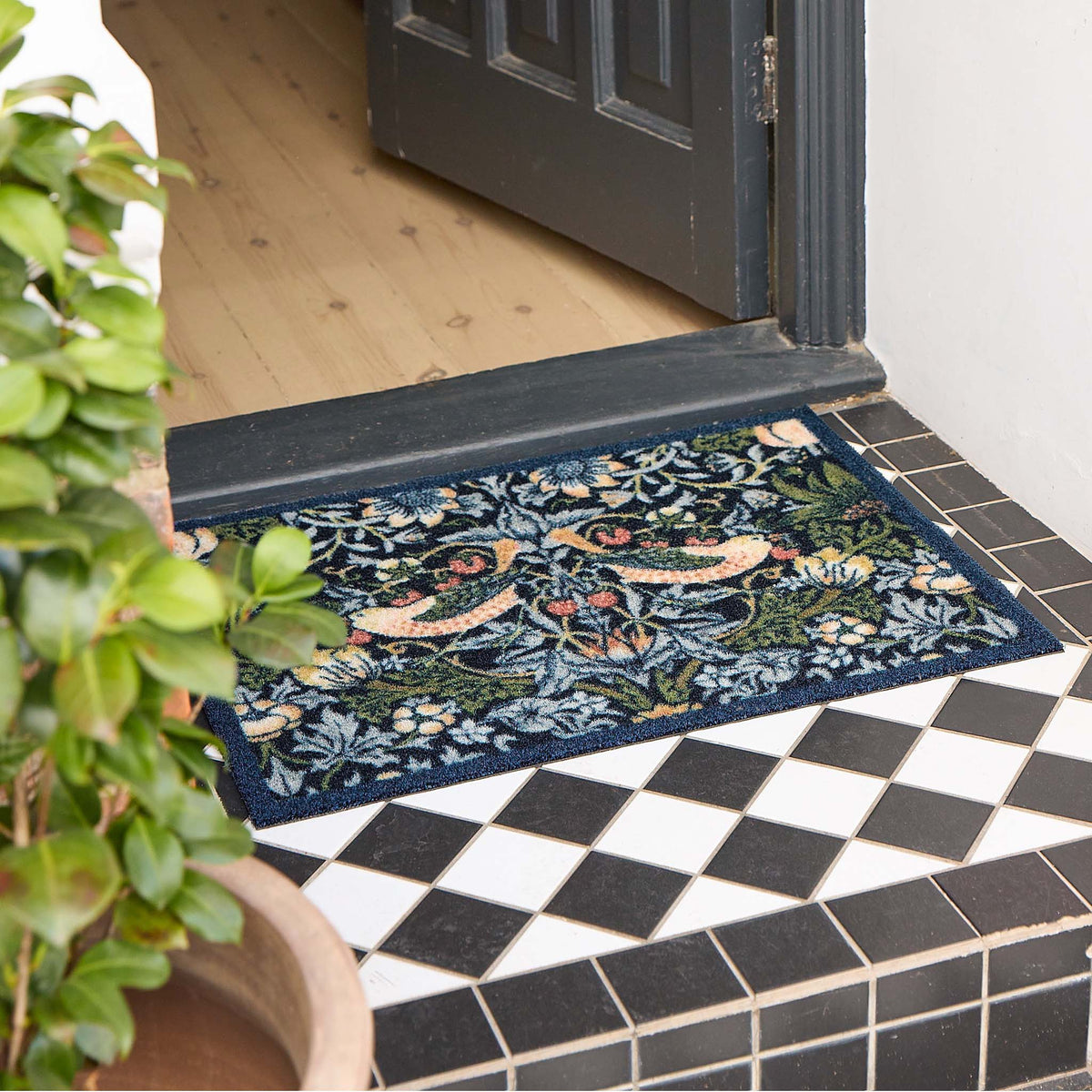 A rectangular outdoor doormat with a dense floral and bird pattern in navy, green, beige, and orange, placed on a black-and-white tiled porch threshold with a wooden door and potted plant visible.