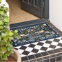 A rectangular outdoor doormat with a dense floral and bird pattern in navy, green, beige, and orange, placed on a black-and-white tiled porch threshold with a wooden door and potted plant visible.
