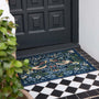 A rectangular outdoor doormat with an intricate floral and bird pattern in navy blue, green, beige, and orange, placed on a black and white tiled porch in front of a dark paneled door.