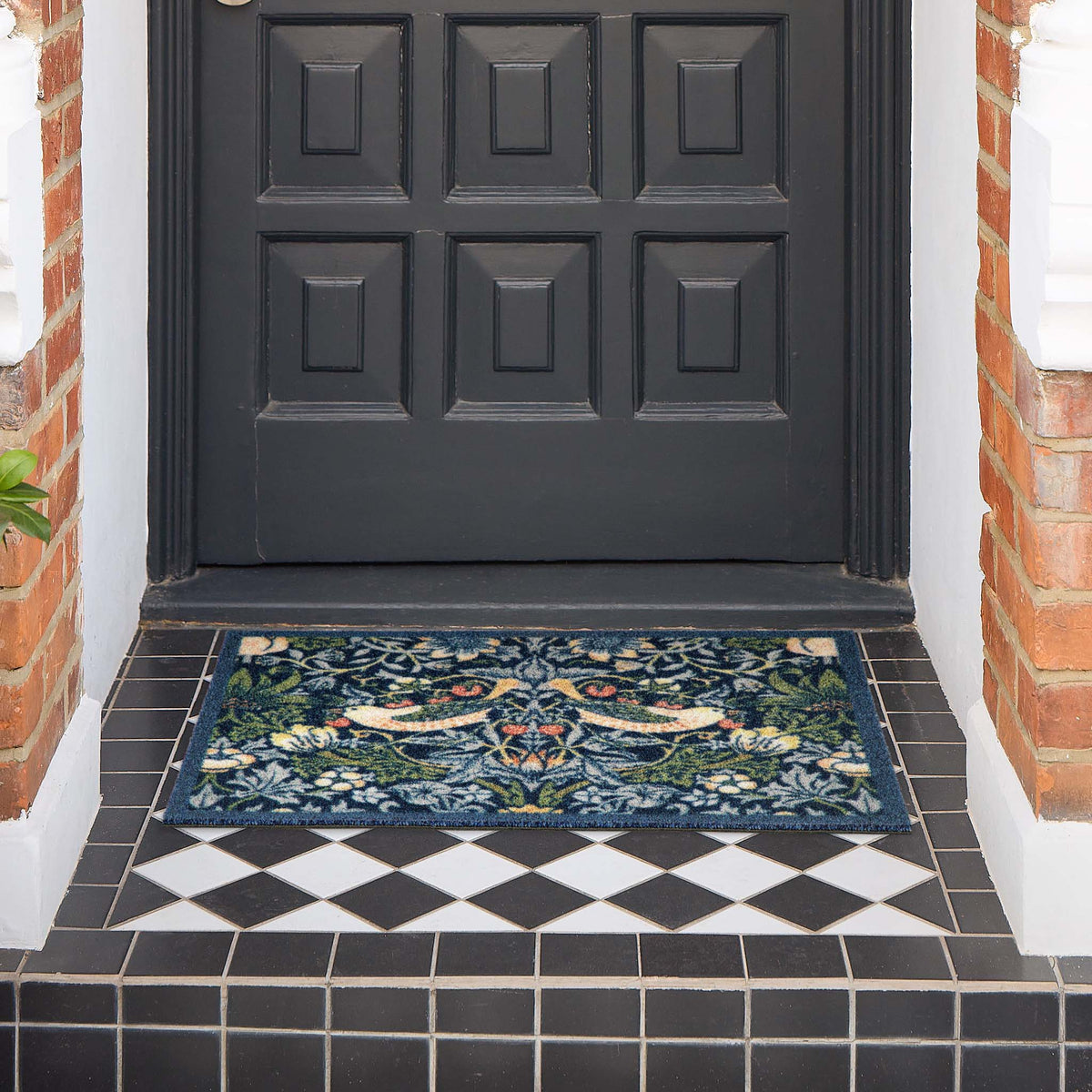 A rectangular outdoor doormat with an intricate floral and bird pattern in navy blue, green, beige, and orange, placed on a black and white tiled porch in front of a dark paneled door.