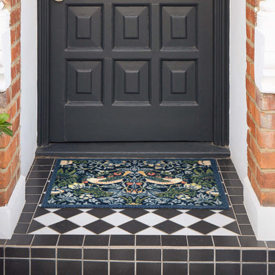 A rectangular outdoor doormat with an intricate floral and bird pattern in navy blue, green, beige, and orange, placed on a black and white tiled porch in front of a dark paneled door.