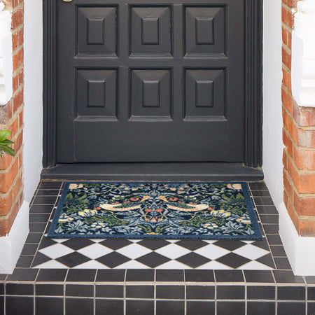 A rectangular outdoor doormat with an intricate floral and bird pattern in navy blue, green, beige, and orange, placed on a black and white tiled porch in front of a dark paneled door.