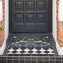 A rectangular outdoor doormat with an intricate floral and bird pattern in navy blue, green, beige, and orange, placed on a black and white tiled porch in front of a dark paneled door.
