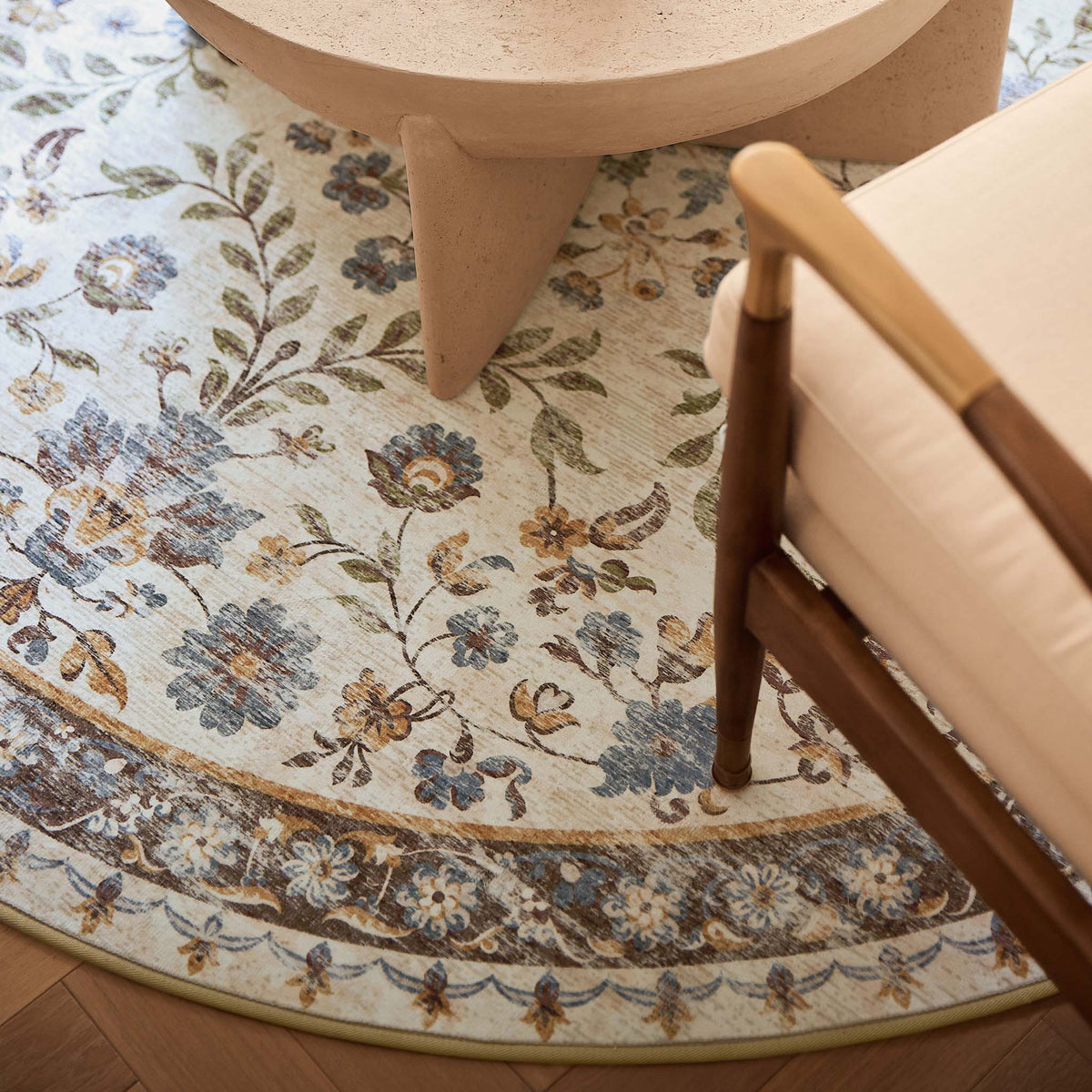 A round area rug with a vintage floral pattern in muted blue, beige, and brown tones, styled in a modern living room setting under a light wood table and next to a wooden armchair.