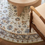 A round area rug with a vintage floral pattern in muted blue, beige, and brown tones, styled in a modern living room setting under a light wood table and next to a wooden armchair.
