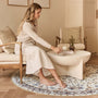 A woman in a beige long-sleeve shirt and white skirt sits barefoot on a round patterned rug in a styled living room, reaching toward a small table on a cream-colored coffee table.