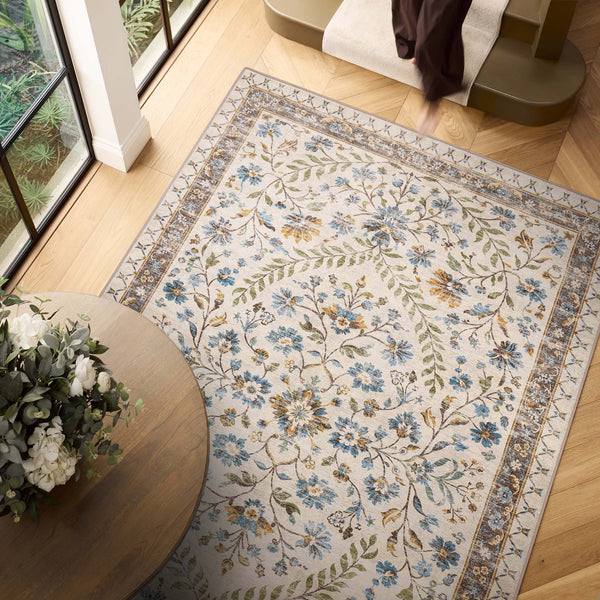 An area rug with a traditional floral pattern in blue, green, and gold on an ivory background, styled in a bright room with light wood floors, a round wooden table with flowers, and a staircase.
