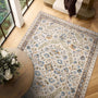 An area rug with a traditional floral pattern in blue, green, and gold on an ivory background, styled in a bright room with light wood floors, a round wooden table with flowers, and a staircase.
