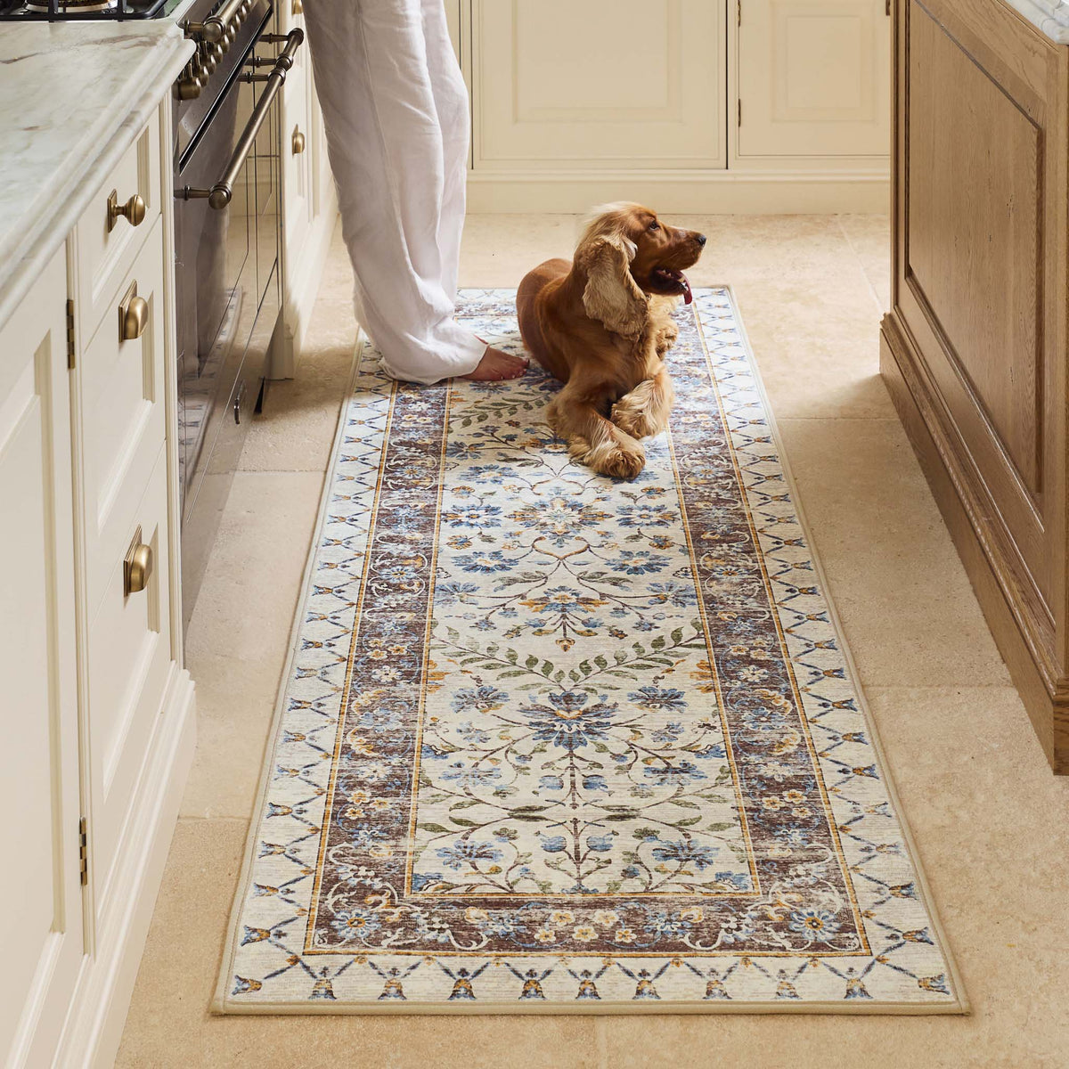 A rectangular area rug with a traditional floral pattern in beige, blue, and brown tones, shown in a kitchen setting with a dog lying on it and a person's legs visible at the top.