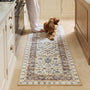 A rectangular area rug with a traditional floral pattern in beige, blue, and brown tones, shown in a kitchen setting with a dog lying on it and a person's legs visible at the top.