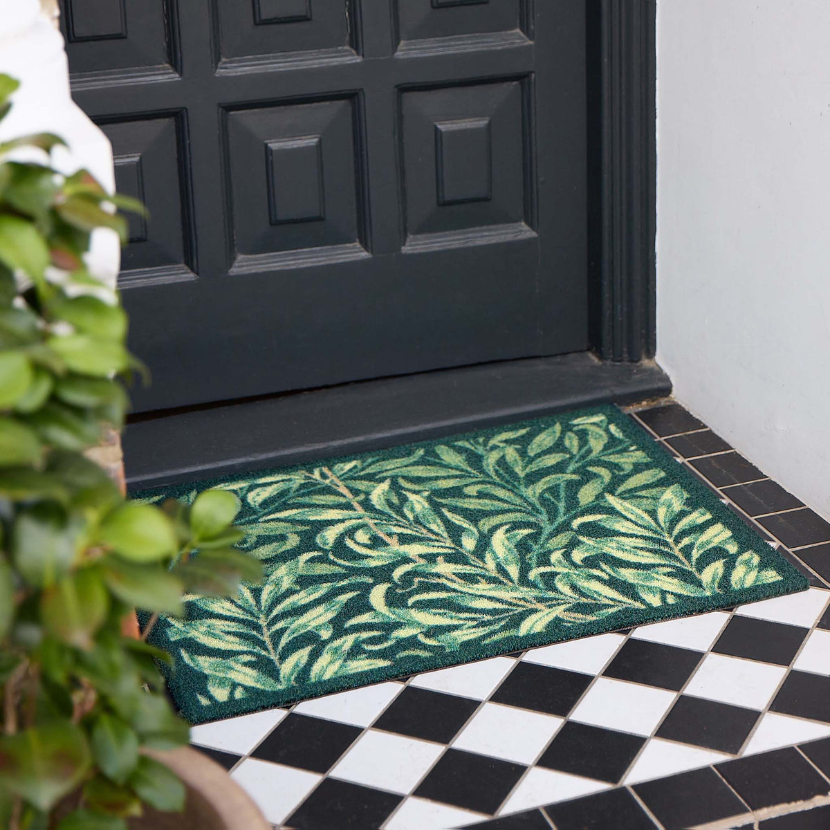 A rectangular green outdoor doormat with a light green palm leaf pattern, placed in front of a black front door on a black and white checkered tile floor, with a potted plant on the left.
