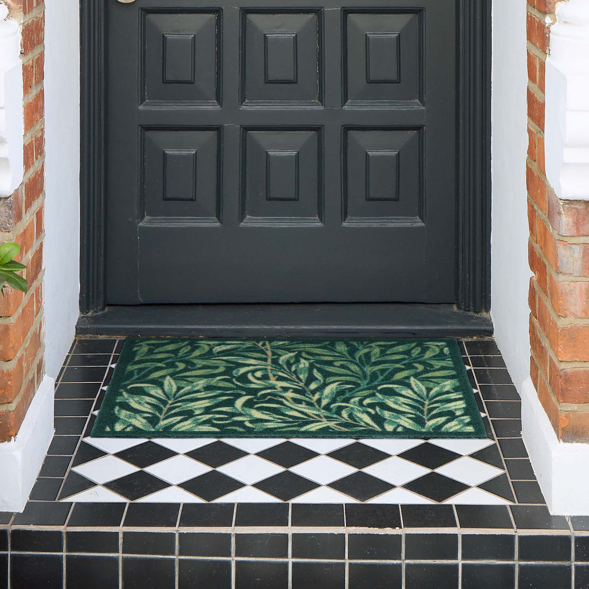 A rectangular green outdoor doormat with a leafy vine pattern, placed in front of a black paneled front door on a tiled porch.