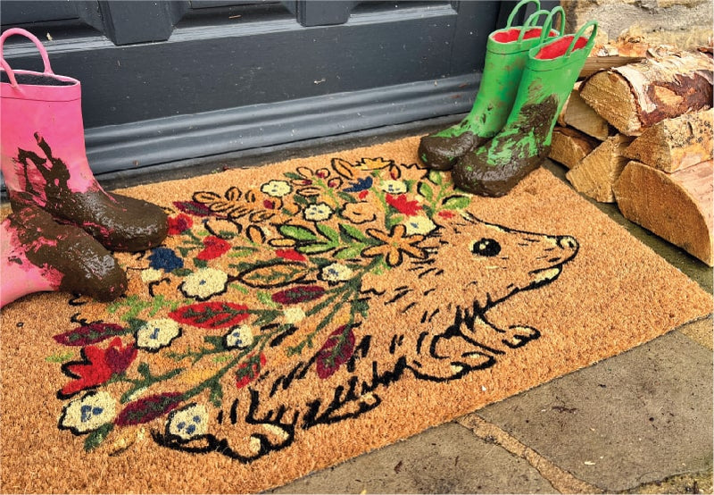 A coir doormat with a floral hedgehog design at a front door with muddy wellies on